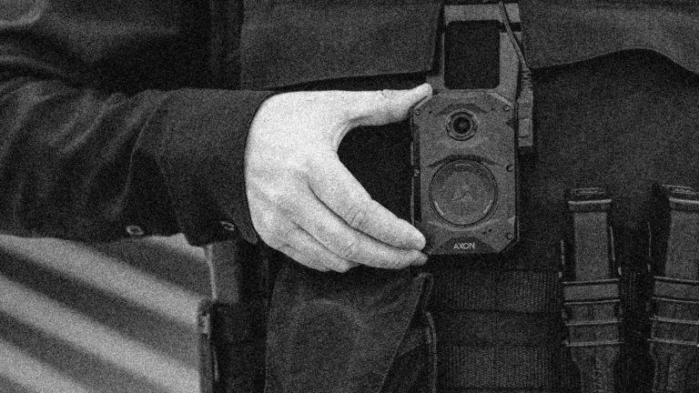 A black and white grainy close-up of a police officer’s hand adjusting an Axon body-worn camera attached to their uniform. The officer’s tactical gear, including straps and equipment pouches, is visible in the background.