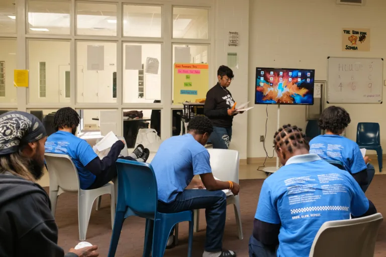 A group of people in blue shirts are seated in a room with a woman standing in front of a large screen displaying an abstract digital background. The setting appears to be educational, with posters and a whiteboard in the background.