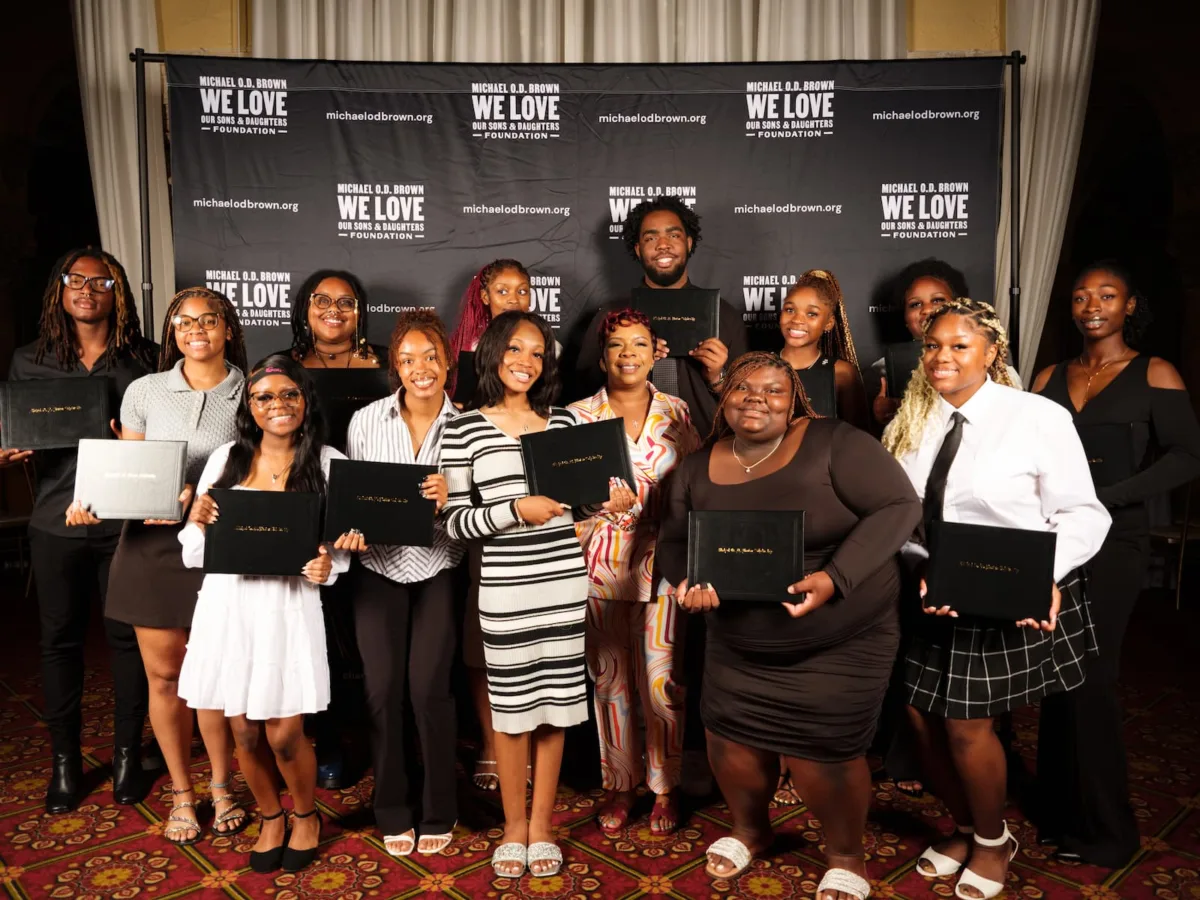A group of people stand together holding certificates in front of a backdrop with the text 'Michael O.D. Brown We Love Our Sons & Daughters Foundation.'