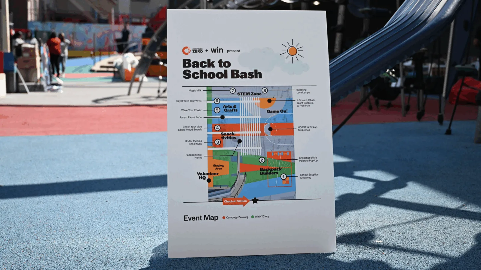 A sign titled "Back to School Bash" with an event map detailing various activities such as Arts & Crafts, STEM Zone, Snacktivities, and Game On, set on a colorful outdoor playground with people in the background.